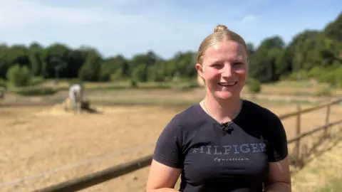 A woman with tied back blonde hair smiles while standing in a field. Behind her is a wooden fence and beyond that is a white horse eating hay.