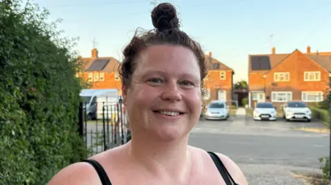 Martin Heath/BBC Kristina Hedley stands outdoors on a residential street in front of a driveway and a row of brick houses. The person has dark hair tied in a high bun and is wearing a black sleeveless top. Several cars are parked in front of the houses, and hedges, gates and utility poles line the street. The image is lit by low evening sunlight.