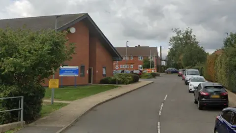 The entrance to Melton Mowbray Hospital, with cars parked on the right and a red brick building with blue entrance sign on the left