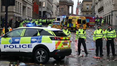 A large group of uniformed police officers cordon off the street with several police and ambulance vehicles on the road, with litter all around and crowds in the distance, in Liverpool on Monday.