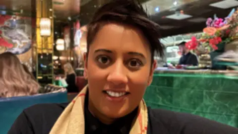 Naveen Jaspal sitting in a restaurant with a bar behind her. She is wearing a black top and a scarf and smiling at the camera.