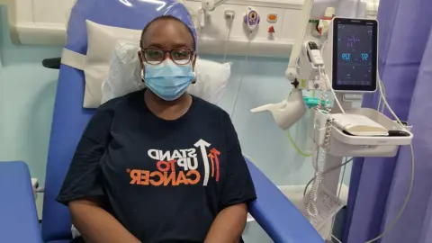 PA Media Simone Boothe wearing a "Stand up to cancer" t-shirt in a hospital, wearing a blue face mask. 