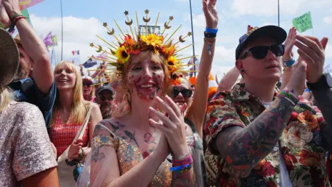 PA Media A woman wearing a flower crown and a glittery dress is clapping while standing in a crowd in front of the Pyramid stage.