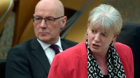 Shona Robison in a red blazer and patterned scarf speaks while standing in the Holyrood chamber, with John Swinney seated individual in the background.