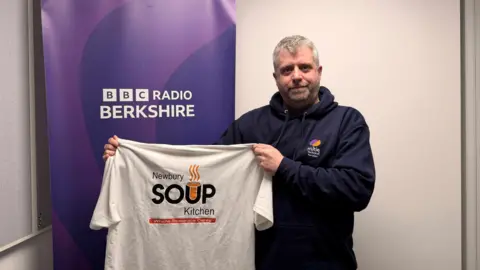 Gavin Weir wearing a navy hoodie holding a white t-shirt which reads Newbury Soup Kitchen with the BBC Radio Berkshire logo in the background.