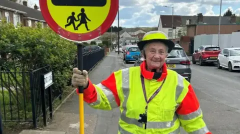 City of Wolverhampton Council An elderly woman in a crossing warden's hi-viz jacket and hat, holding the lollipop sign. She stands on a pavement next to a road with lots of cars parked on it.