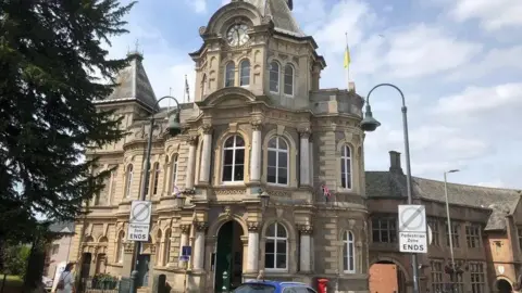 Tiverton Town Hall. A tall building with large windows and a clock in the top. There are lampposts and "pedestrian zone ends" signs. To the left is a tree with green leaves. The sky is blue with white clouds. 