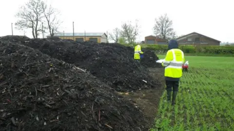 Environment Agency officers in high-vis jackets standing in a field next to a pile of compost.