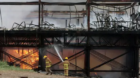 PA Media Two firefighters tackle the fire on Fleetwood pier after the blaze destroyed most of the historic pier. One is holding a hose to the burned out shell and one walks past with his head down past an area under the pier that is still ablaze