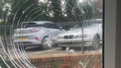 Charlie Williams A broken window with cracks in the glass forming a circular pattern and two cars seen through the glass