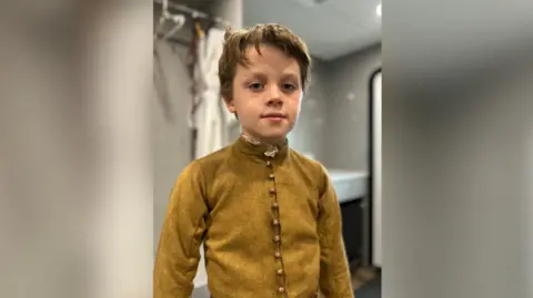 James Lintern, 11, looks at the camera. He has dark hair and a cowlick. He wears a mustard coloured Tudor-period button down doublet. Other costumes can be seen hanging in the background. 