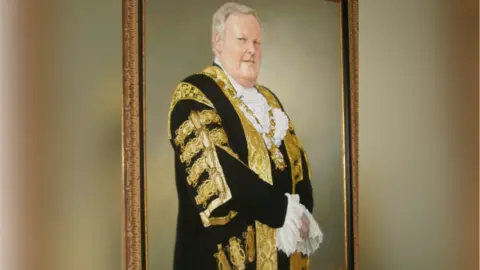 Belfast City Council The portrait of Lord Browne is hanging on a wall. He's wearing an official suit. It's black with gold designs and ruffles are peeping out of his sleeves and top.