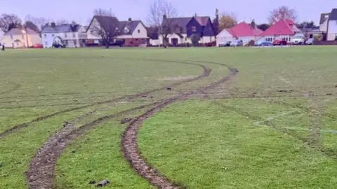 Alex Jones Long lines of tire tracks left by a car that has repeatedly driven over the green. They are widespread and criss-cross in certain areas.