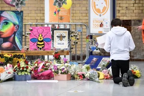 LightRocket/Getty Images A young man kneels in front of the floral memorial at Victoria station during the commemoration. Colourful flowers, posters and pictures of Manchester bee icons are shown.