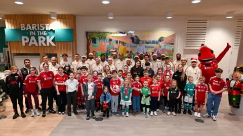 Alder Hey Children's Hospital Patients wearing Liverpool kits are smiling as they stand in front of the smiling Liverpool squad members in a large room, with a Christmas tree and a large nutcracker figurine at either side of them.
