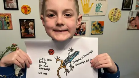 A young boy smiling, holding a hand drawn picture of a lizard