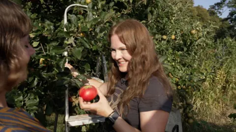 A woman with long auburn hair and a grey t-shirt holds a red apple standing next to a ladder and apple trees.