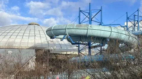 BBC A derelict building with a domed roof and a rusting slide. A grey fence and thin trees surround the building. There is a curved slide that was once part of the swimming pool section