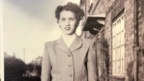 Tony Lester A black and white photo of a woman with short curly hair, wearing a suit jacket. She stands on a street with, to her left, a house with a latticed window. Behind her look to be further homes in the distance.