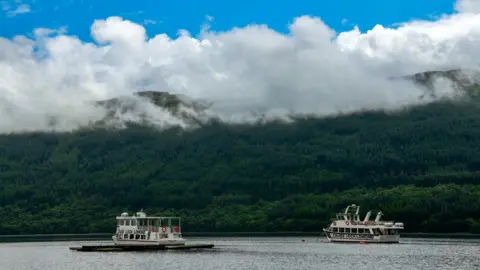 John Lang Two boats float on the water with dense forests on a hill at the other side of the shore. A white cloud hangs low and blue skies can be seen above the clouds.