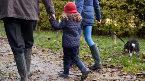 A child wearing a blue coat and red hat with face turned away from the camera walks between two adults