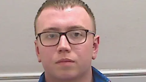 A police mugshot of Cameron Miller. He is a young man with short light brown hair and black rimmed glasses.