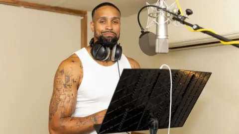 BBC Studios A picture of Ashley Banjo in a recording studio with a white vest on and headphones around his neck