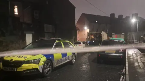 A street with police cordon, police car and other vehicles parked.