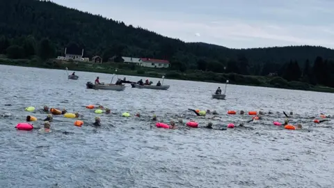 Roz Rapp A crowd of people swimming in a river with support boats floating alongside them.