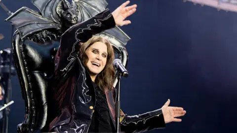 A man sits on a black throne with a bat motif at the top. He has shoulder-length brown hair and he is wearing black. There is a microphone in front of him.