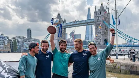 Stefano Capella Five men standing in front of London's tower bridge, holding a trophy. They are all wearing blue tops and shorts