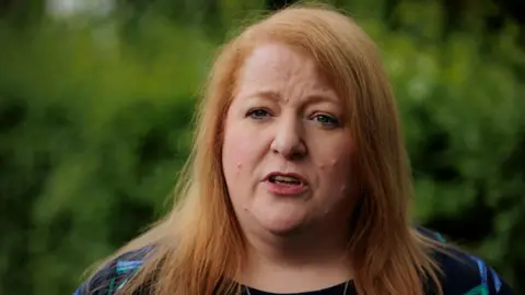 Naomi Long stands outside. She has ginger hair and is wearing a navy top with a blue and green pattern. There are trees behind her.