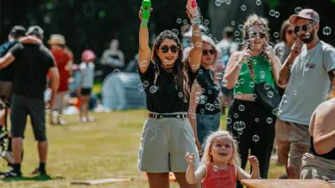 Digbeth Dining Club A woman in sunglasses holds up two bubble blasters and a small girl with facepaint on reaches up to the bubbles in the sunshine.