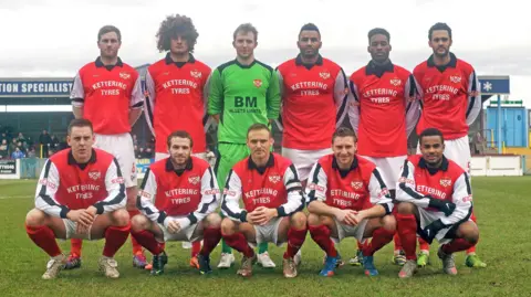 Peter Short/@northantslegend on X A Kettering Town team photo, consisting of 11 players either squatting or standing on the pitch together. They are wearing a red shirt with white sleeves, a black collar and "Kettering Tyres" written in white in the middle. Their shorts are white and their socks are red.