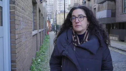 Francesca Albanese stands outside on a London road. She wears a black coat and beige patterned scarf. She has glasses and dark, curly hair.