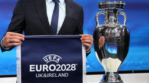 A blue Euro 2028 UK and Ireland pennant being held up alongside the European Championship trophy