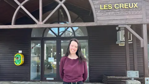 Jennifer Heald pictured outside Les Creux pavilion. She is wearing a plum coloured top and and is smiling. She has dark brown hair. In the background, the entrance to Les Creux is visible. The pavilion is mostly made of wood. 