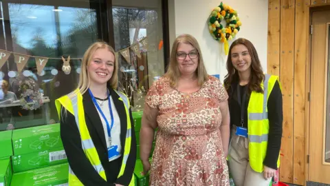 A photo of three women smiling into the camera. Two women on the sides have high visibility jackets on. The woman in the middle has a flowery dress on. They are all smiling into the camera. 
