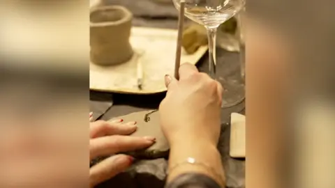 A pair of hands using a wooden tool to make marks in a piece of clay. A wine glass sits on the table