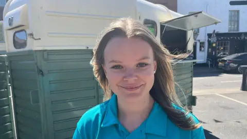 John Devine/BBC Lydia Cowling is smiling at the camera. She is standing in front of a green and cream horse box. She has long, fair hair and is wearing a blue top.