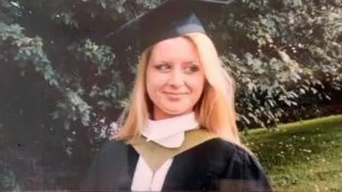 FAMILY HANDOUT Jo in hat and gown at a graduation ceremony. She is standing outside, in front of a tree.