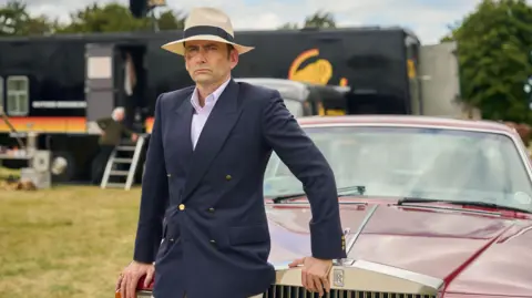 PA Media David Tennant as Tony Baddingham. He is lent against a Rolls Royce classic car and is wearing a navy-blue double breasted blazer. He is wearing a cream boat hat. 