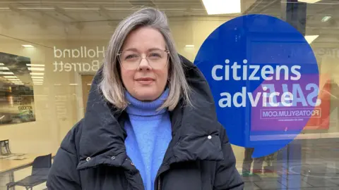 Sarah McPhie. She has shoulder length grey hair, glasses, dark jacket and blue top. She is stood in front of the Stockton and District Advice and Information Service glass window with the blue and white Citizen's Advice logo on it.