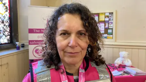 Night Angels CEO Chrissie Lowry is wearing a pink Hi-vis jacket and has shoulder-length brown curly hair. She is looking into the camera and smiling.