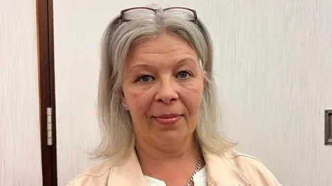 A woman wearing a cream-coloured jacket and white top is looking directly at the camera. She has grey hair and is wearing a pair of glasses on top of her head. She is standing against a light-coloured door.