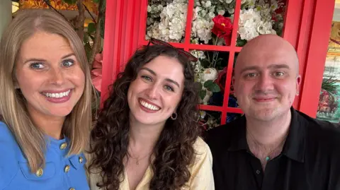 The Tudor Trio Three people all smiling and standing in front of a red phone box which has been filled with flowers. From left, they are Nicola Tallis, with blonde shoulder-length hair and wearing a blue top with gold buttons; Kate McCaffrey, with long curling dark brown hair over a yellow shirt; and Owen Emmerson who is waring a black open neck shirt. 