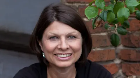 Theatre founder Joanna Carrick has short dark hair. She is smiling at the camera. An earring in her right ear is visible. Behind her is a brick wall and some foliage.