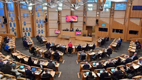 Getty Images Finance Secretary Shona Robison (standing in the centre wearing a red jacket) delivers the Scottish Budget for 2026-2027 in the chamber of the Scottish Parliament in Edinburgh on 13 January, 2026. A number of MSPs can be seen at wooden desks with computer screens. 