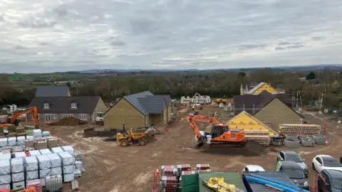 An aerial view of a building site next to countryside and hills in Somerset. Large bags of building materials are visible as well as machinery including an orange digger