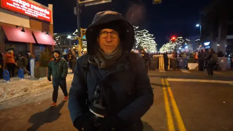 Tom Bateman wearing a hooded coat near protests on a blocked off street in Minneapolis